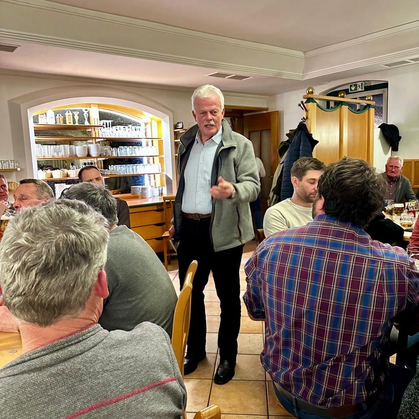 Ein älterer Mann in einem grauen Mantel steht und spricht zu einer Gruppe von Menschen, die an einem Tisch in einem Restaurant sitzen. Der Raum hat weiße Wände und eine Holztheke mit Regalen.