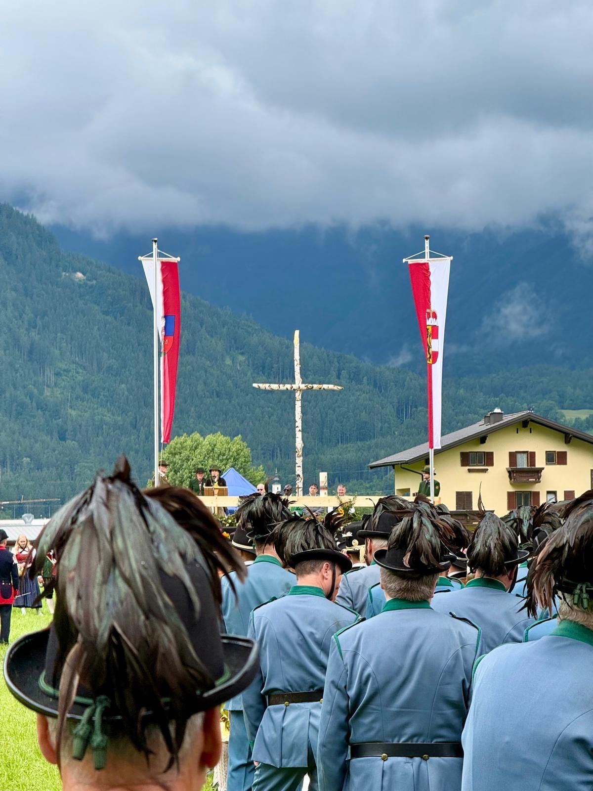 Eine Gruppe von Menschen in traditionellen Uniformen mit Federhüten steht vor einem hölzernen Kreuz vor einer Bergkulisse. Hinter ihnen befinden sich Fahnen mit roten und weißen Streifen, und ein gelbes Haus ist im Hintergrund sichtbar.