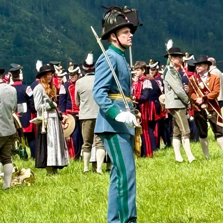 Eine Gruppe von Menschen in historischen Uniformen steht auf einer Wiese, mit Bergen im Hintergrund. Einige halten Instrumente, und ein Mann hält ein Schwert.