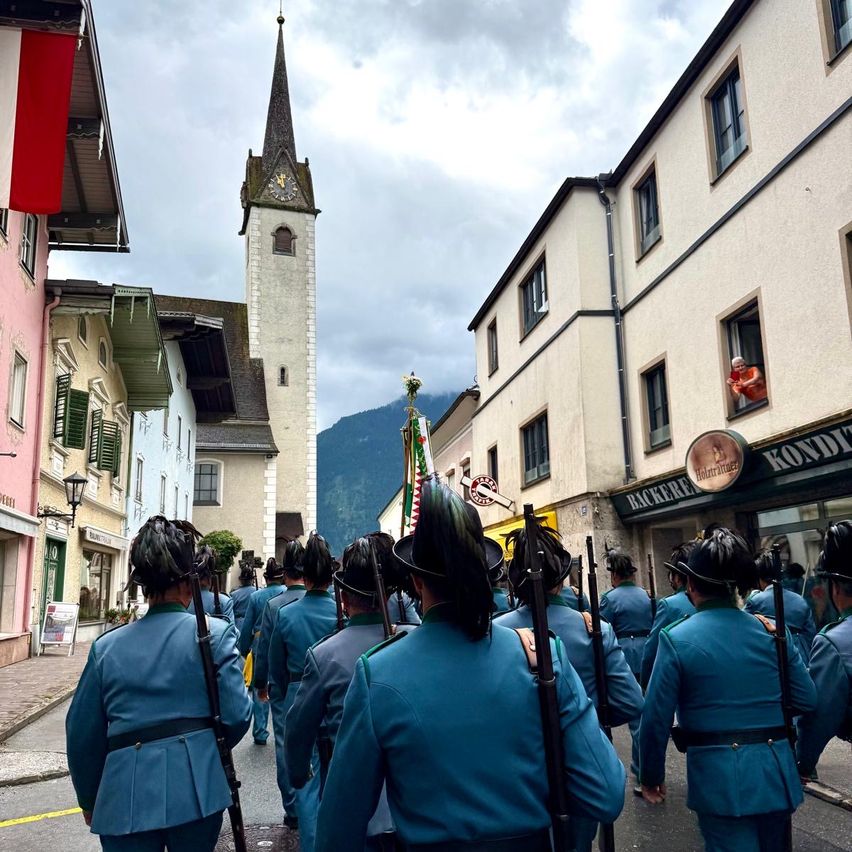 Eine Marschkapelle in blauen Uniformen zieht mit Gebäuden und einem Turm im Hintergrund die Straße entlang.