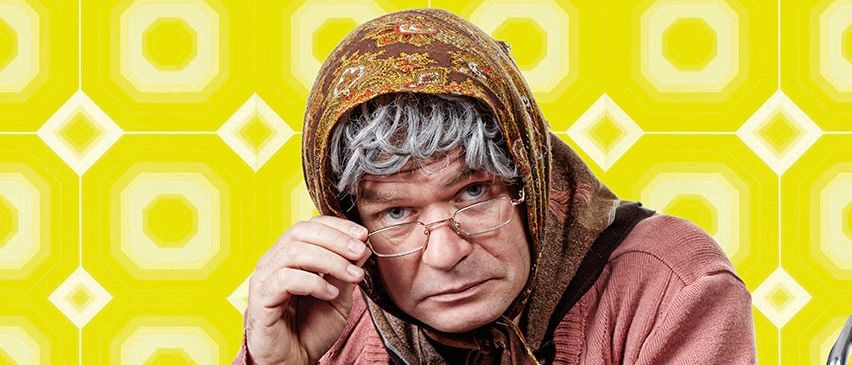 Eine ältere Frau im Rollstuhl mit Brille und Schal sitzt vor einem gelben Hintergrund mit einer Tüte Brezeln.