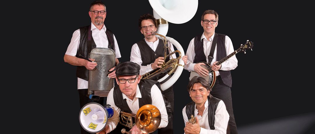 Fünf Musiker posieren für ein Foto vor schwarzem Hintergrund. Sie halten Musikinstrumente wie ein Posaune, ein Banjo und eine Tuba.