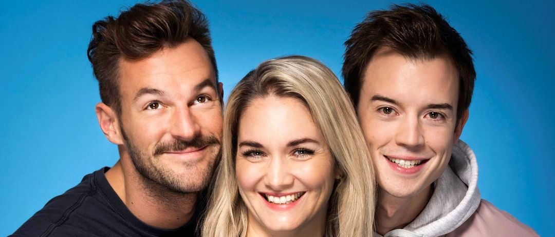 Drei Personen, zwei Männer und eine Frau, posieren für ein Foto. Der Mann links trägt ein schwarzes ärmelloses Shirt, die Frau in der Mitte eine schwarze Bluse und der Mann rechts eine rosa Kapuzenjacke. Sie alle lächeln in die Kamera.