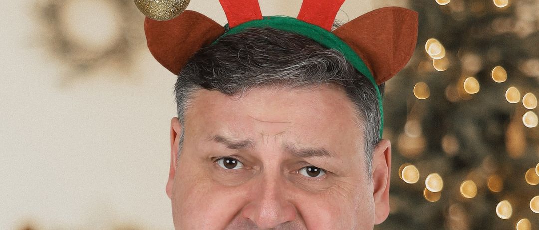 Ein Mann mit weißem Hemd und Rentier-Kopfband steht vor einem Weihnachtsbaum mit verschwommenen Lichtern.