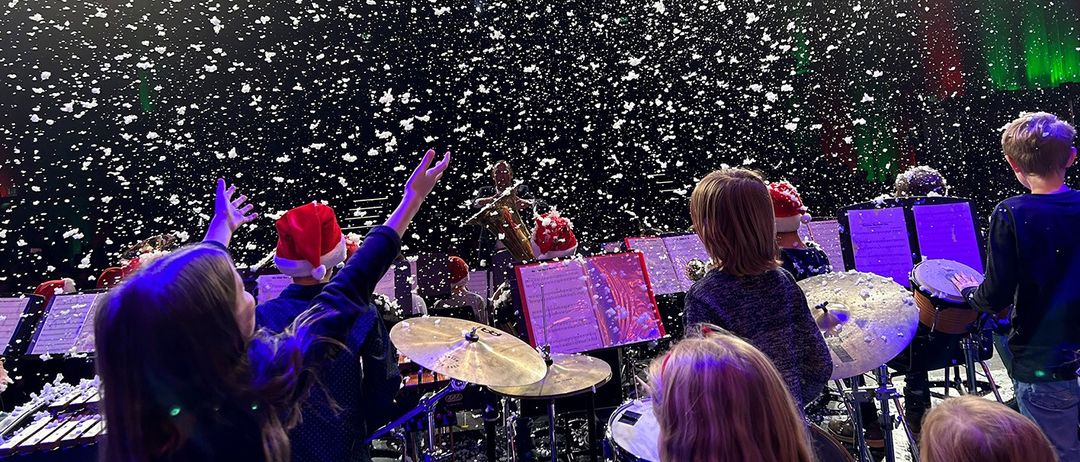 Eine Gruppe von Kindern in Weihnachtsmützen spielt auf der Bühne mit Musikinstrumenten, während künstlicher Schnee von der Decke fällt.