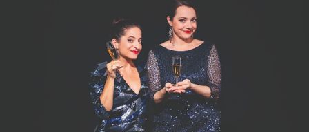 Zwei Frauen in Abendkleidern halten Champagnerflöten vor einem dunklen Hintergrund. Sie lächeln und scheinen zu feiern.