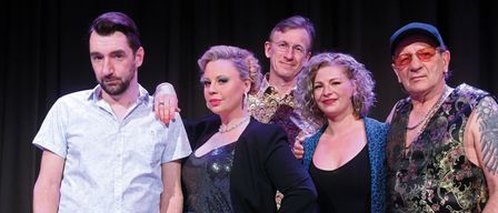 Fünf Menschen stehen eng beieinander und lächeln. Ein Mann links in einem weißen Hemd und Jeans. Die zentrale Figur ist eine Frau in einem schwarzen Oberteil und einer blauen Rock. Hinter ihr steht ein Mann mit Brille und einem bunten Hemd. Zwei Frauen stehen neben ihm, beide tragen blaue Jacken.