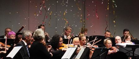 Ein Orchester spielt auf der Bühne, ein Mann steht vor ihnen. Die Musiker spielen ihre Celli, Violinen und Flöten. Das Publikum beobachtet sie. Auf der Bühne stehen auch Blumen.