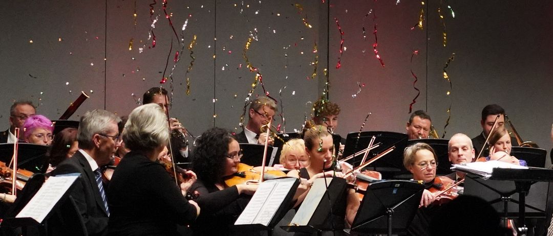 Ein Dirigent steht vor einem Orchester auf der Bühne, Konfetti fällt. Musiker spielen Violinen, Celli und Flöten. Das Publikum schaut aus dem Dunkeln.