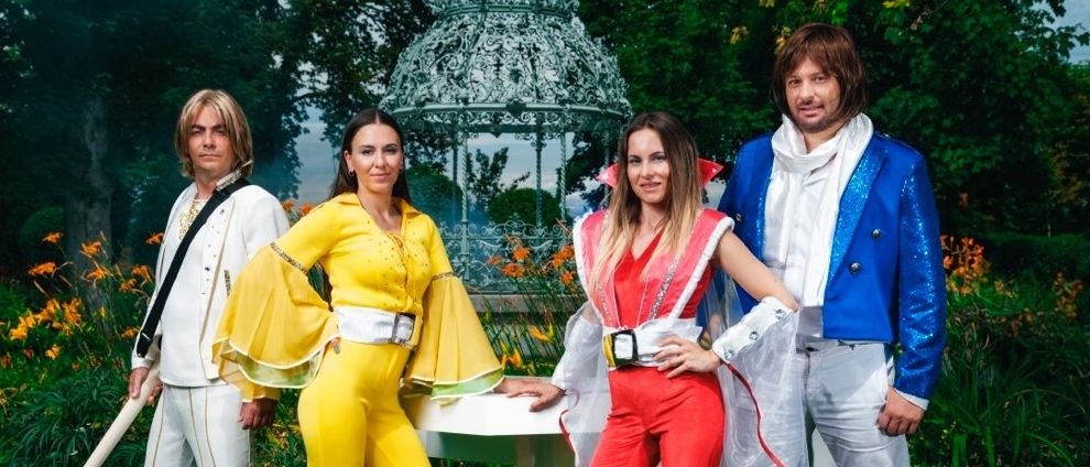 Vier Menschen posieren für ein Foto in einem Garten mit einem Pavillon. Ein Mann hält eine Gitarre, während die anderen farbenfrohe Kostüme tragen.