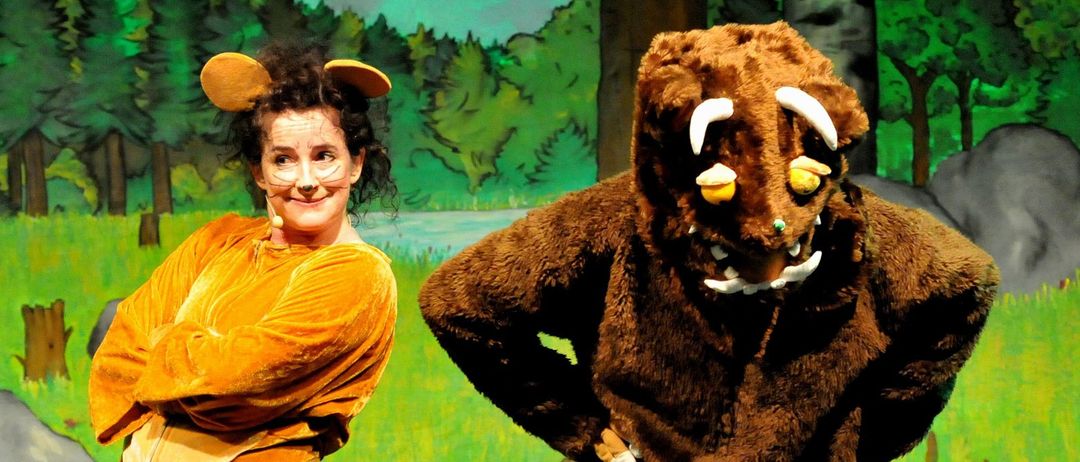 Eine Frau in einem Bärenkostüm steht neben einer Person in einem braunen Monsterkostüm auf der Bühne, vor einer Kulisse eines Waldes.