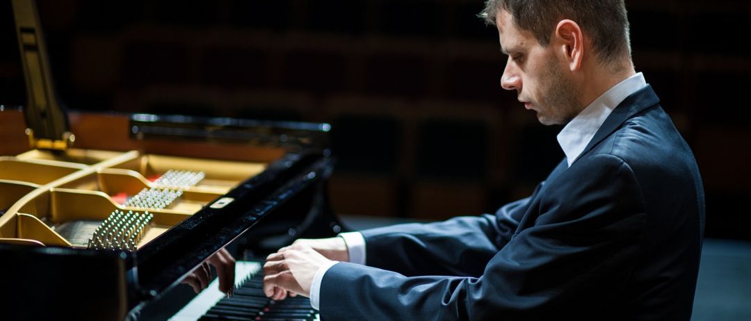 Ein Mann in einem Anzug spielt Klavier in einem schwach beleuchteten Raum. Das Klavier ist schwarz mit goldenen Akzenten. Der Mann konzentriert sich auf sein Spielen und sitzt am Klavier.