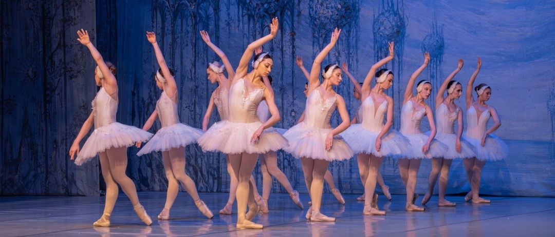 Eine Ballettaufführung mit weiblichen Tänzerinnen in weißen Tütüs, die anmutig auf einer Bühne mit blauen Kulissen auftreten.