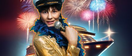 Eine Frau in einem goldglänzenden Kostüm mit Matrosenmütze hält ein Mikrofon mit Feuerwerk im Hintergrund. Ein Kreuzfahrtschiff liegt im Wasser.