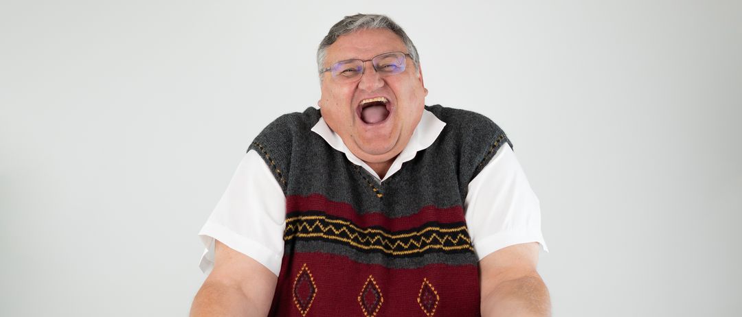 Ein Mann in einem gemusterten Weste und Brille, lacht mit offenem Mund, mit ausgestreckten Armen vor einem schlichten weißen Hintergrund.