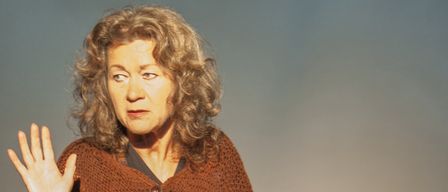 An older woman with curly hair, wearing a brown cardigan, appears to be waving while looking towards the camera.