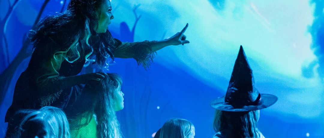 Eine Frau, die als Hexe verkleidet ist, zeigt auf eine Gruppe junger Mädchen, die um ein Feuer auf einer Bühne mit blauem und grünem Hintergrund sitzen.