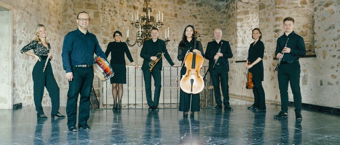 Eine Gruppe von Musikern posiert in einem Raum mit Steinwänden, einer Kronleuchter und einer Treppe. Sie halten verschiedene Instrumente.