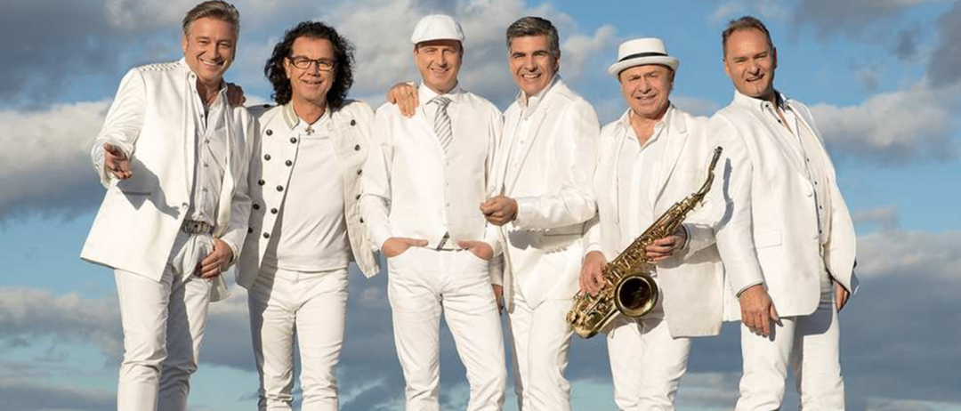 Sechs Männer in passenden weißen Anzügen stehen auf einem Grasfeld und posieren für ein Foto. Ein Mann hält eine Saxophon.