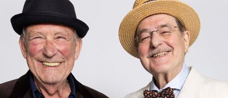 Zwei ältere Männer lächeln und stehen eng beieinander. Der Mann links trägt einen schwarzen Blazer über einem blauen Hemd. Der Mann rechts trägt einen weißen Anzug, einen Hut und eine Fliege.