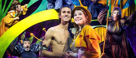 Eine Bühnenaufführung mit zwei Personen vorne, die lächeln. Ein Mann in einem Lendenschurz und eine Frau in einem gelben Kleid. Dahinter sind auch andere Darsteller auf der Bühne.