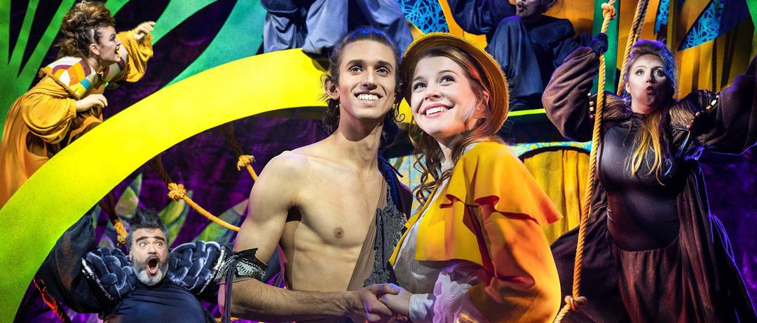 Eine Bühnenaufführung mit zwei Personen vorne, die lächeln. Ein Mann in einem Lendenschurz und eine Frau in einem gelben Kleid. Dahinter sind auch andere Darsteller auf der Bühne.