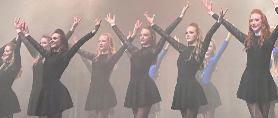 Eine Gruppe weiblicher Tänzerinnen tritt auf der Bühne auf, mit erhobenen Armen und lächelnden Gesichtern, unter rauchiger Wirkung und Bühnenlicht.