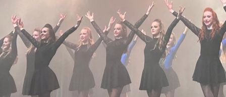 Eine Gruppe weiblicher Tänzerinnen tritt auf der Bühne auf, mit erhobenen Armen und lächelnden Gesichtern, unter rauchiger Wirkung und Bühnenlicht.