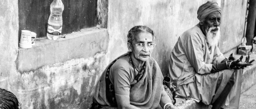 Ein Schwarz-Weiß-Foto von drei älteren Menschen, die auf einer Mauer sitzen. In der Nähe befinden sich Teller, Tassen und Flaschen. Die Frau links trägt einen Armreif. Der Mann rechts trägt einen Turban.