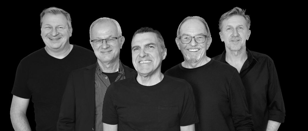 Fünf Männer, alle lächelnd, stehen zusammen in einem Schwarz-Weiß-Foto. Sie tragen lässige Kleidung, einschließlich Brillen und Uhren.