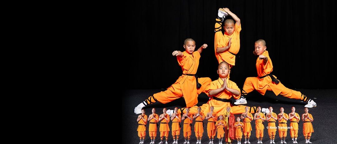 Eine Gruppe von Kampfkünstlern in orangefarbenen Uniformen tritt in einer Reihe auf der Bühne auf.
