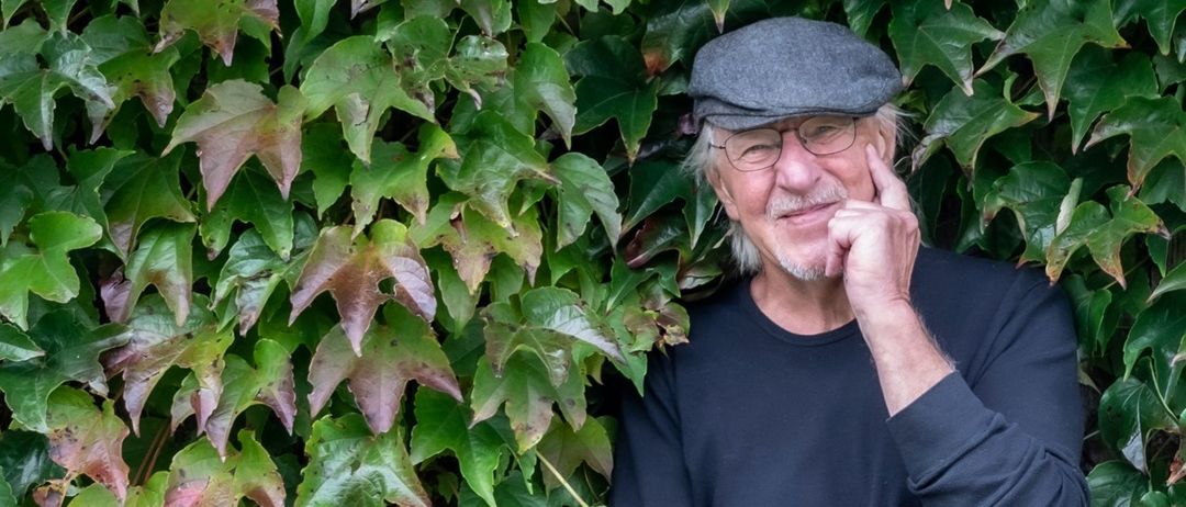 Ein älterer Mann mit Brille und Hut steht neben einer grünen Wand. Er lächelt und berührt sein Kinn mit der Hand.