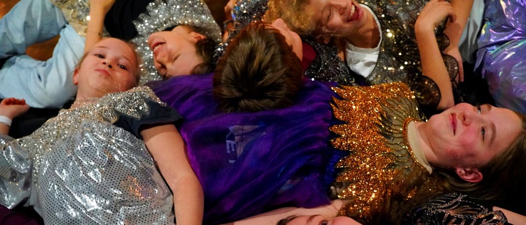Mehrere junge Menschen in funkelnden Kostümen liegen auf dem Boden. Einige lächeln, andere scheinen zu lachen. Die Kostüme schimmern mit Pailletten.