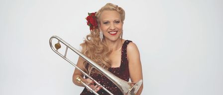 Eine lächelnde Frau in einem roten Paillettenkleid mit einer roten Blume im Haar posiert mit einem Posaune.