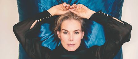 Eine Frau mit kurzen Haaren liegt auf einem blauen Hintergrund und trägt ein schwarzes Oberteil mit Knopfdetails. Sie hält ihren Kopf mit beiden Händen und schaut nach vorne.