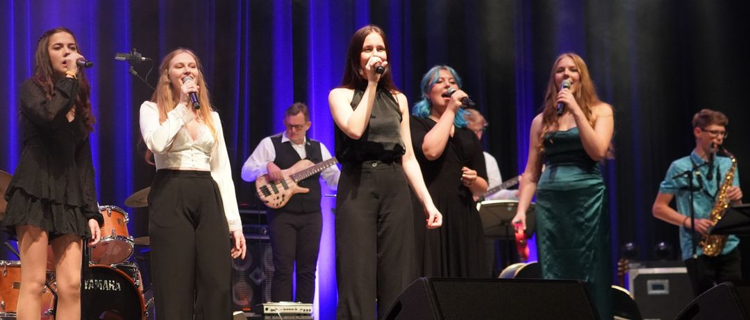 Eine Band tritt auf der Bühne mit einer weiblichen Sängerin vorn auf. Die Band besteht aus einem Gitarristen und einem Schlagzeuger. Die Bühne ist mit lila Beleuchtung erhellt.