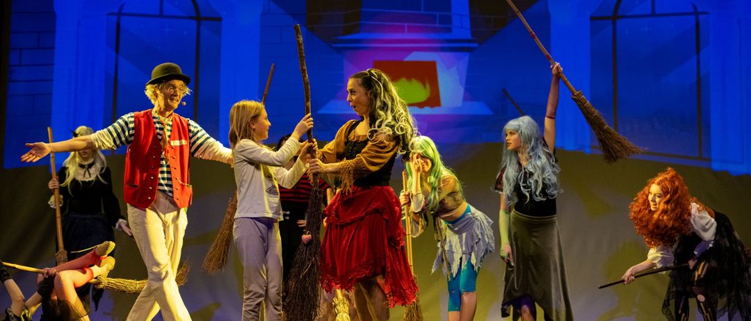 Eine Gruppe von Kindern und Erwachsenen steht auf der Bühne, in Kostümen gekleidet. Sie halten Besen in der Hand, wobei ein Mädchen in einem roten Rock einen Stock hält. Sie scheinen in einem Theaterstück aufzutreten.