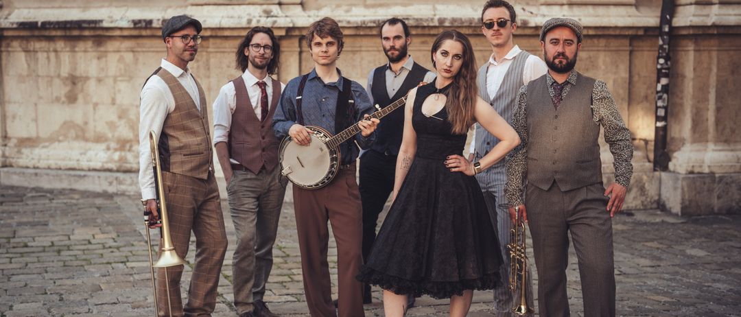 Eine Gruppe von Musikern in Vintage-Kleidung steht vor einem Gebäude. Eine Frau in einem schwarzen Kleid hält eine Banane, während ein Mann eine Trompete spielt.