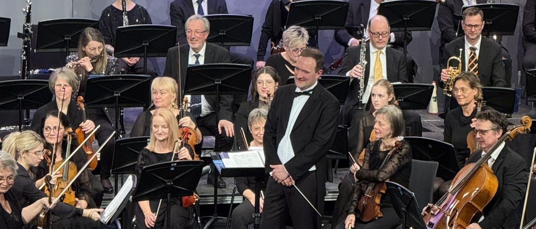Ein Symphonieorchester auf der Bühne, mit Musikern, die verschiedene Instrumente spielen. Ein Dirigent steht vorne und leitet die Aufführung. Die Bühne ist mit bunten Blumen geschmückt.