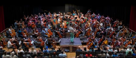 A large ensemble of cellists, including adults and children, perform on stage in a well-lit concert hall with an audience seated in rows. A conductor stands on a small platform in front of them.