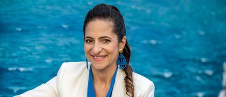 Eine lächelnde Frau in einem weißen Blazer und einem blauen Schal posiert neben einem Pool. Sie trägt blaue Ohrringe.