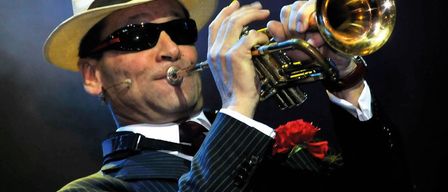 Ein Mann in einem Anzug spielt eine Trompete auf der Bühne unter einem Scheinwerfer. Er trägt einen Fedora, Sonnenbrille und eine rote Rose.