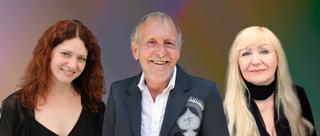 Drei Menschen, zwei Frauen und ein Mann, stehen lächelnd vor einem bunten Hintergrund. Der Mann trägt einen schwarzen Anzug mit einem silbernen Pik-Design.