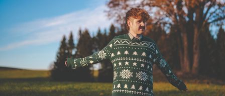 Ein Mann in einem Weihnachtspullover steht in einem Feld mit Bäumen im Hintergrund.