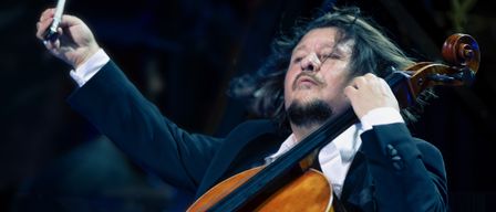 Ein Mann mit langen Haaren und Bart spielt Cello bei einem Konzert, hält einen Bogen in der linken Hand. Er trägt einen dunklen Anzug.