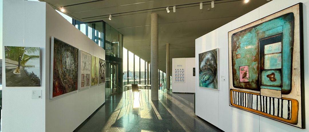 Eine Kunstgalerie mit modernen Gemälden, die an weißen Wänden ausgestellt sind. Der Raum hat eine hohe Decke mit Lichtern und einem langen Korridor. Es gibt Glaswände, die das Außengelände zeigen.