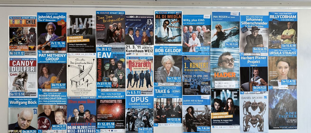 Eine Wand mit mehreren Postern. Verschiedene Veranstaltungen und Bands werden beworben, darunter Dee Dee Bridgewater, Al Di Meola und Bob Geldof. Daten und Uhrzeiten sind aufgeführt.
