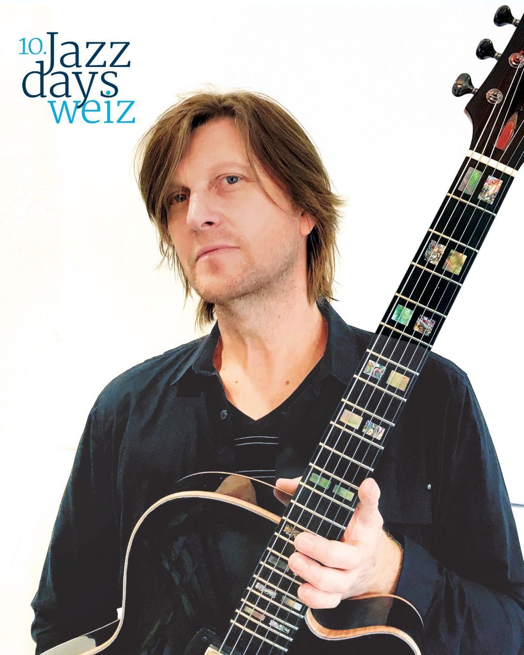 Ein Mann mit langen Haaren und einem schwarzen Hemd hält eine Gitarre mit einem einzigartigen Design, das farbige Quadrate auf dem Griffbrett aufweist, vor einem weißen Hintergrund.