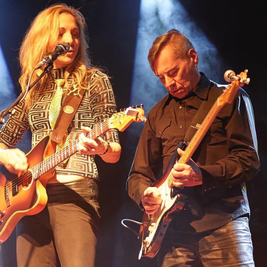 Eine Frau und ein Mann spielen Gitarren auf der Bühne, beleuchtet von Scheinwerfern. Die Frau singt in ein Mikrofon, während der Mann nach unten schaut.