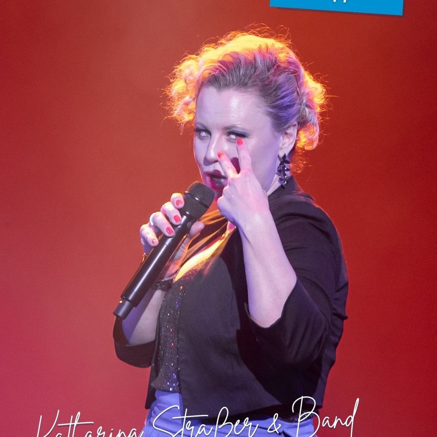 Eine Frau singt in ein Mikrofon auf der Bühne. Sie hat roten Nagellack und ihre Augen sind geschlossen. Sie trägt ein schwarzes Oberteil. Die Bühne hat einen roten Hintergrund. Das Bild hat einen Wasserzeichen.